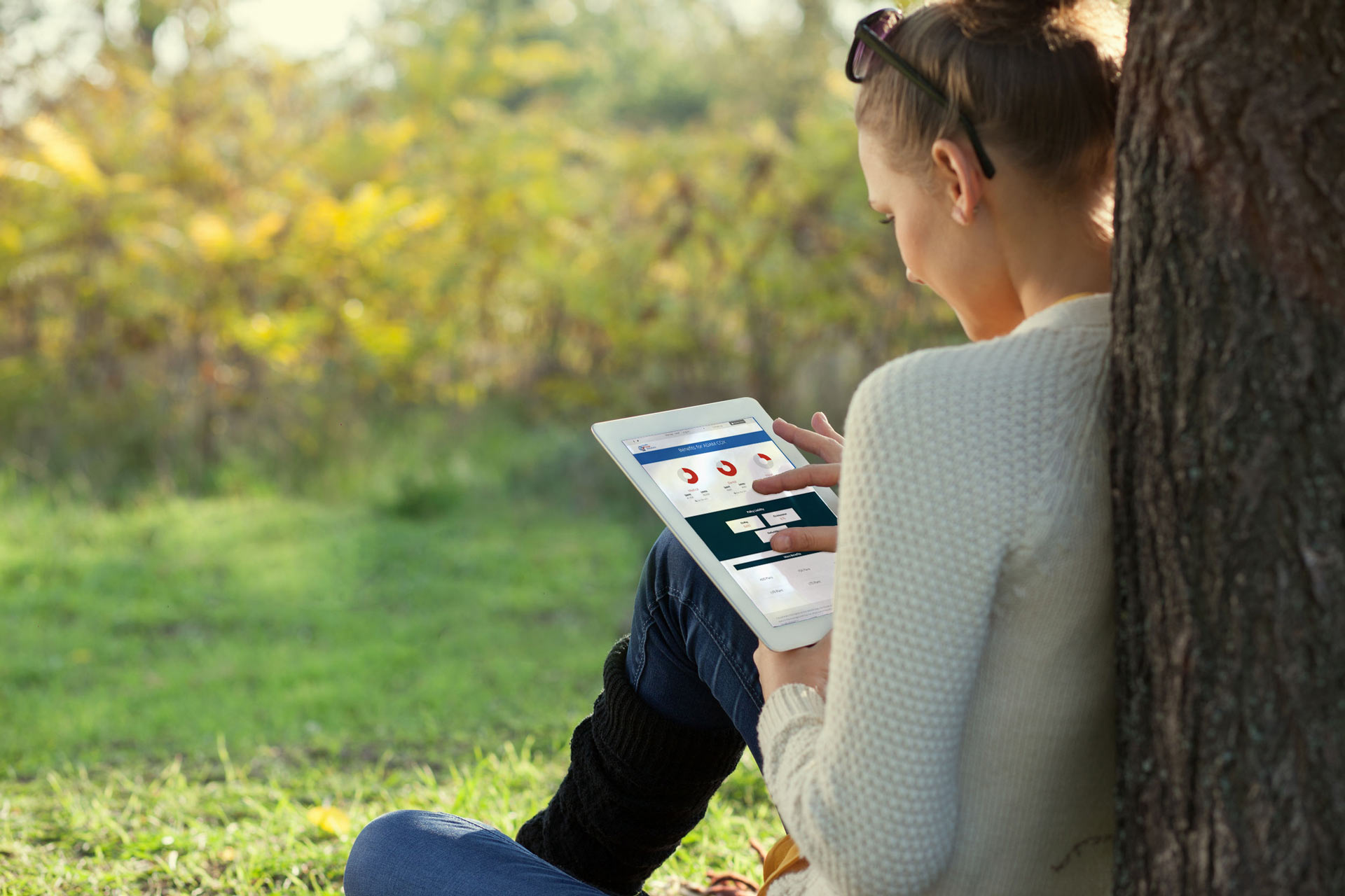 woman sitting on the grass under a tree with using an ipad to look at health plan benefit charts