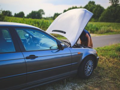 person looking under the hood of a car on the side of a country road