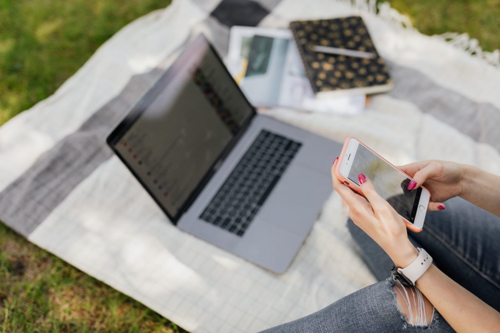 woman holding cell phone while sitting on a picnic blanket on the grass, next to a laptop and notebooks