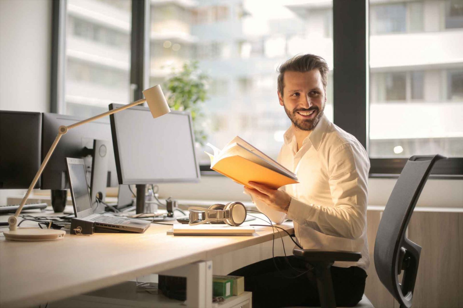 Smiling man sitting at computer desk while reading a book