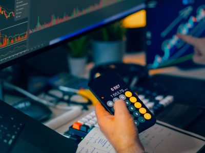 hand holding a calculator and another hand pointing at a screen with a line graph